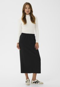 Jeune femme portant un haut blanc à manches longues, une jupe midi noire et des baskets blanches avec un motif noir, se tenant devant un fond blanc uni.