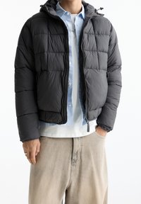 Schwarze gesteppte Steppjacke mit Kapuze, ausgestattet mit einem Frontreißverschluss und kontrastierenden Nähten. Getragen über einem hellblauen Hemd mit Knöpfen.