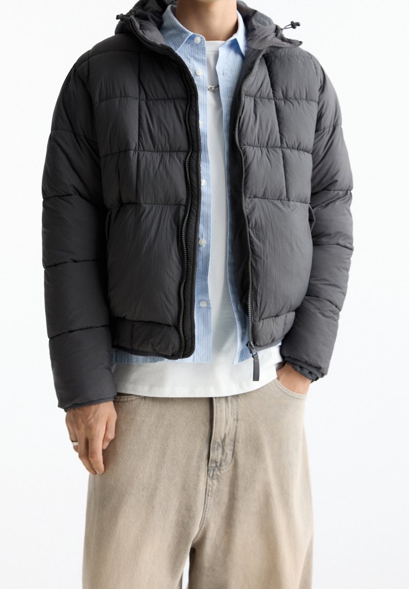 Schwarze gesteppte Steppjacke mit Kapuze, ausgestattet mit einem Frontreißverschluss und kontrastierenden Nähten. Getragen über einem hellblauen Hemd mit Knöpfen.