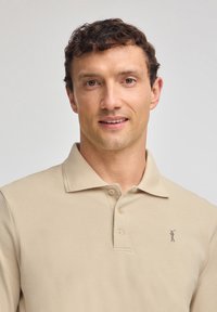 Beżowa koszulka polo z kołnierzykiem i trzema guzikami, wykonana z fakturowanej tkaniny. Posiada haftowane logo golfisty po lewej stronie klatki piersiowej.
