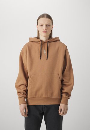Brown hoodie made of soft fabric, featuring a front pocket, drawstrings, and logo detail on the chest. Paired with dark jeans.