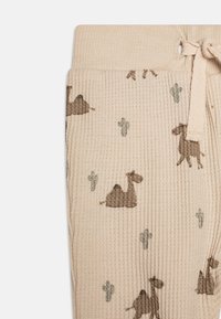 Beige ribbad tyg med ett lekfullt mönster av kamel och kaktusar i mörkbrunt och grönt, med dragsko i midjan för justerbar passform.