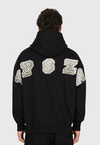 Mann in schwarzem Kapuzenpullover mit großen reflektierenden Buchstaben "AP SZA" über dem oberen Rücken und den Schultern, von hinten auf einem schlichten Hintergrund abgebildet.