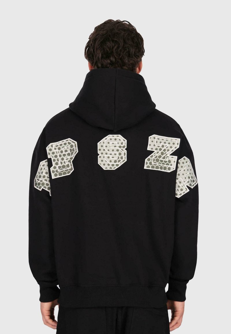 Mann in schwarzem Kapuzenpullover mit großen reflektierenden Buchstaben "AP SZA" über dem oberen Rücken und den Schultern, von hinten auf einem schlichten Hintergrund abgebildet.