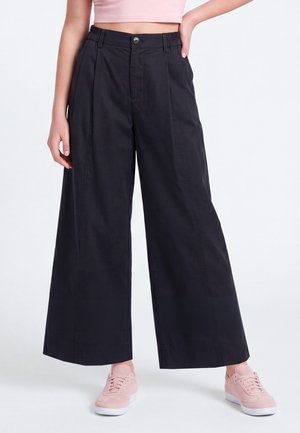 Person iført sorte vidde bukser med høj talje, lyserød cropped top og lyserøde sneakers, står med den ene hånd på hoften.