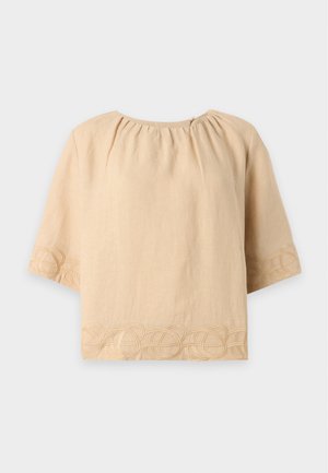 Kvinde iført en løs beige top med broderi, brede beige bukser, sorte sko og holder en sort håndtaske mod en ensfarvet hvid baggrund.