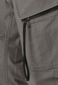 Γκρι ύφασμα με λεία υφή διαθέτει τσέπη με φερμουάρ και μαύρο κορδόνι, τονίζοντας πρακτικές σχεδιαστικές λεπτομέρειες.