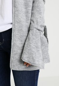 Szary sweter z dzianiny, z szerokimi rękawami, z detalem w postaci wiązania. Miękka tekstura. Połączony z białą bluzką i ciemnoniebieskimi dżinsami.