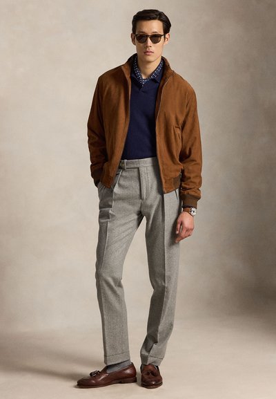 Man draagt een bruine suède jas, een marineblauwe trui, een sjaal met stippen, grijze broek, bruine leren loafers, zonnebril en een polshorloge, staand met handen in de zakken.