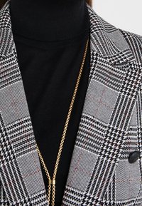 Col roulé noir sous un blazer pied-de-poule noir et blanc avec un long collier en chaîne dorée pendu à l'avant.