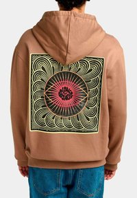 Sudadera marrón con un gran diseño gráfico en la parte posterior, que presenta líneas en espiral y un motivo central de sol circular rojo y negro.