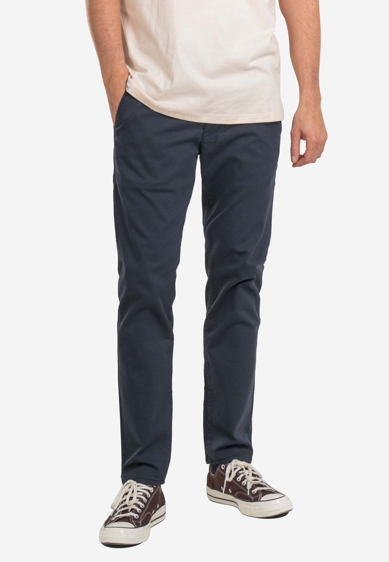 Pantalones de algodón chino azul marino con corte slim, frente plano y bolsillos estándar. Combinados con zapatillas marrones y una camiseta de color claro.