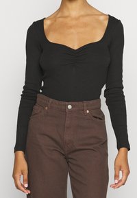Donna che indossa una maglietta nera a maniche lunghe con scollo arricciato e pantaloni marroni a vita alta, in piedi con le braccia rilassate lungo i fianchi.