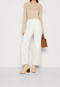 Beige gerippter Langarmoberteil, kombiniert mit weißen Jeans mit hohem Bund und weitem Bein. Das Model hält eine kleine braune Lederhandtasche. Die Schuhe sind beige Stiefeletten mit Absatz.