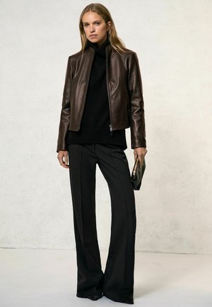 Mujer de pie con una chaqueta de cuero marrón oscuro sobre un suéter de cuello alto negro, pantalones negros de pierna ancha y sosteniendo un bolso de mano negro.