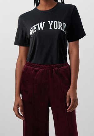 Personne portant un t-shirt noir avec le texte "NEW YORK" et un pantalon en velours bordeaux, debout devant un fond clair uni.