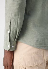 Cămașă verde din in cu mâneci lungi, nasturi albi la manșete, material ușor texturat. Purtați-o peste un material bej cu un buzunar tip patch.