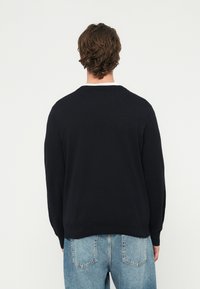 Mężczyzna stojący tyłem, ubrany w czarny sweter z długim rękawem i jasnoniebieskie dżinsy, na jednolitym białym tle.