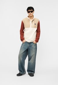 Νεαρός άνδρας που φοράει μαύρα γυαλιά ηλίου, φλις πουλόβερ με μπεζ και σκουριά χρώματα, φαρδιά μπλε τζιν και μαύρα αθλητικά παπούτσια, στέκεται με τα χέρια στις τσέπες.