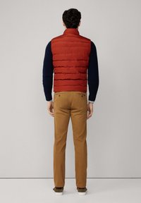 Chaleco acolchado rojo con un cuello alto, llevado sobre un suéter azul marino, combinado con pantalones caqui y zapatos marrones, mostrando un atuendo casual desde atrás.