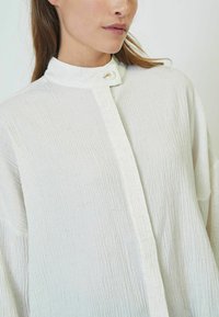 White button-up shirt with a high collar, made of textured fabric featuring subtle crinkles and a single button closure at the neck.