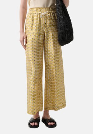 Person wearing yellow wide-leg patterned pants with white drawstring, black sandals, beige top, holding a large black woven bag.