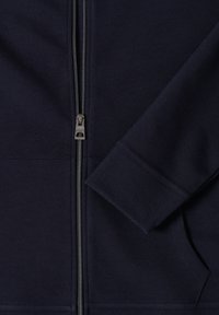 Gros plan d'une veste zippée bleu marine foncé montrant la fermeture éclair, le poignet de manche et une poche avant avec des coutures visibles.