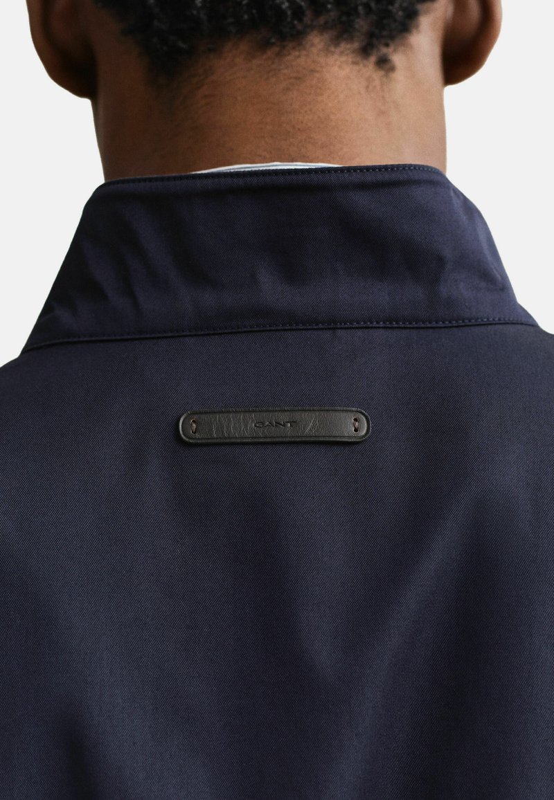 Vue de dos d'une personne portant une veste bleu marine à col montant avec une petite étiquette rectangulaire en cuir cousue dans le haut du dos.