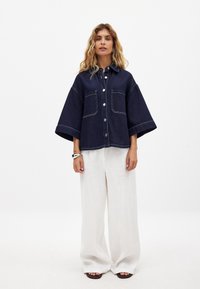 Kvinde med bølget blondt hår iført en mørkeblå oversized skjortejakke, brede hvide bukser, sorte sandaler og et sølvarmbånd.
