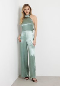 Mėtų žalia satino jumpsuit'as su aukštu halter tipo kaklu, plačiomis kojomis ir švelniu blizgesiu, derinamas su juodais aukštakulniais bateliais su dirželiais.