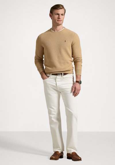 Beige gebreide trui met ronde hals, witte jeans en bruine leren schoenen. Gedetailleerd met een logo op de borst en een gestructureerde stof.