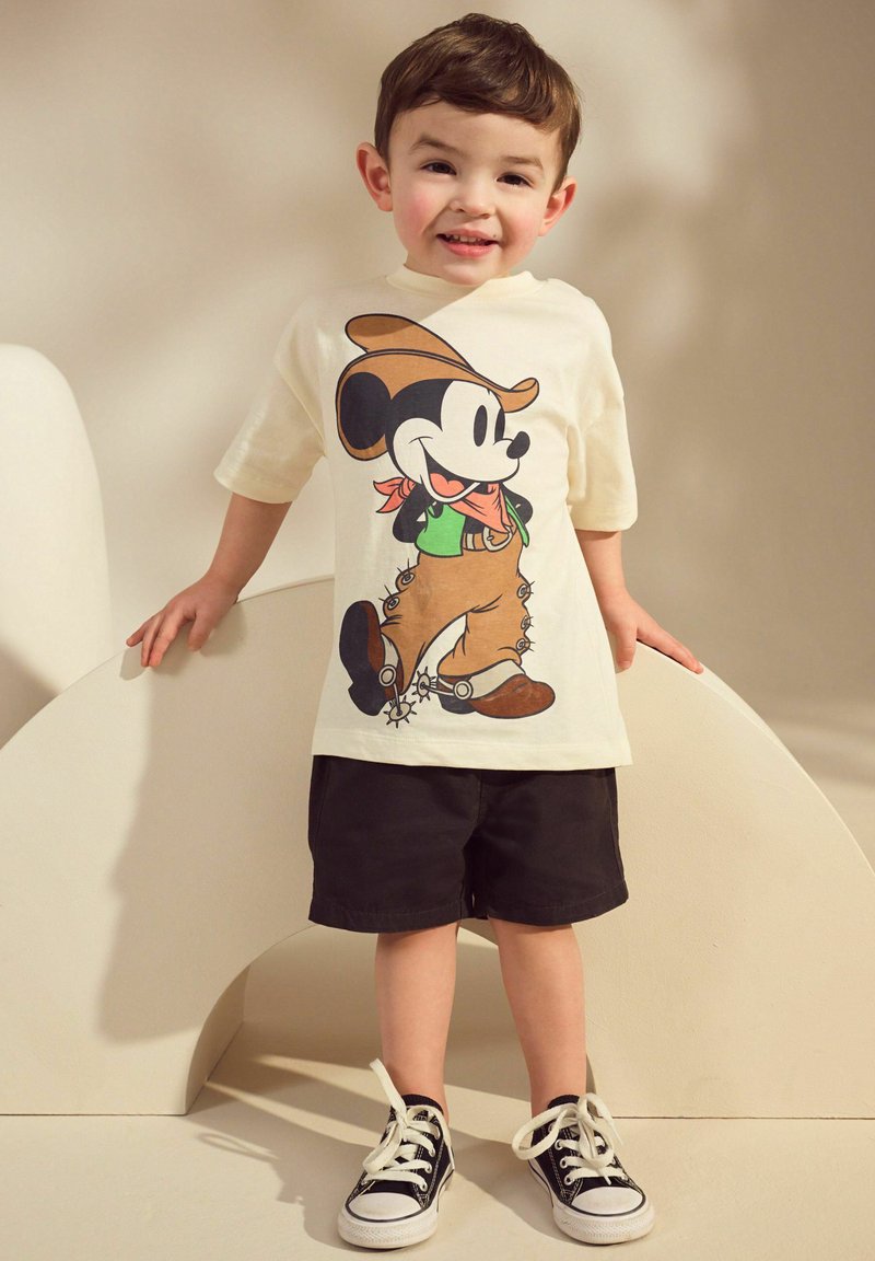 Hvid t-shirt med et farverigt Mickey Mouse grafik, parret med sorte shorts og sorte sneakers med hvide snørebånd. Barn står på en lys baggrund.