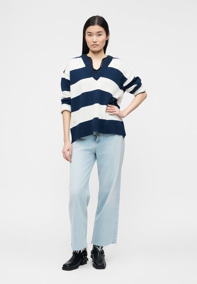 Νεαρή γυναίκα που φοράει μια φαρδιά μπλε-ναυτικό και λευκή ριγέ πουλόβερ, ανοιχτόχρωμα παντελόνια με φαρδιά μπατζάκια και μαύρα μποτάκια αστραγάλου, στέκεται με το χέρι στη μέση.