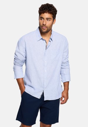 Hombre con camisa de manga larga de color azul claro con las mangas remangadas y pantalones cortos azul marino, de pie con una mano en el bolsillo.