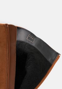 Botín de ante marrón con un forro interior de cuero negro y una etiqueta de marca. Textura suave con cierre de cremallera en un lado.