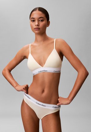 Frau trägt beige Calvin Klein Bralette und passende Unterwäsche mit Logo-Bund, steht mit den Händen in der Hüfte vor grauem Hintergrund.