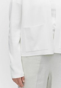 Witte gebreide cardigan met lange mouwen en twee voorzakken, in combinatie met lichtgekleurde linnenbroeken die een soepele textuur laten zien.