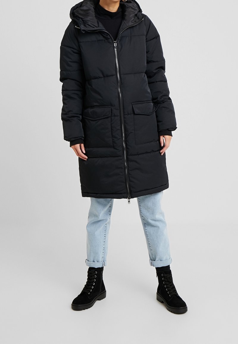 Personne portant un long manteau noir matelassé avec une fermeture éclair à l'avant, deux poches à rabat, un jean bleu clair retroussé aux chevilles, et des bottes noires à lacets.