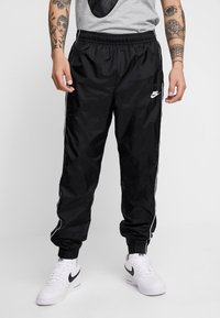 Persona ģērbusies melnās Nike sporta biksēs ar baltām svītrām un baltās Nike kedās, stāv pretī vienkāršai gaišai fona.
