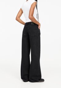 Personne portant un jean noir taille haute et à pattes d'éléphant, un haut sans manches blanc, les cheveux tressés, debout avec les mains dans les poches arrière sur un fond blanc.