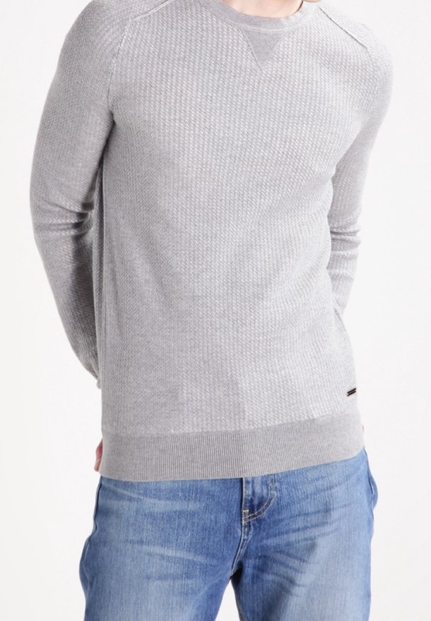 Homme vêtu d'un pull gris clair texturé et d'un jean bleu, debout les mains dans le dos devant un fond uni.