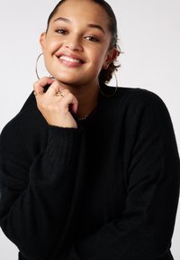 Glunderende vrouw met ringoorbellen en ringen, gekleed in een zwarte trui, met haar kin op haar hand rustend tegen een effen lichte achtergrond.