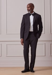 Smoking nero con revers a punta in satin, camicia bianca, papillon nero e scarpe nere lucide, su sfondo neutro.