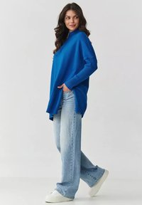 Niebieski oversize'owy sweter dzianinowy z prążkowanymi mankietami, bocznymi szczelinami i wysokim kołnierzem, w połączeniu z jasnoniebieskimi spodniami typu straight-leg i białymi sneakersami.