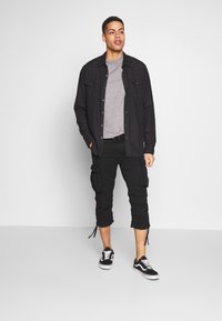 Zwarte button-up shirt over een grijs T-shirt, zwarte cargoshorts en zwarte sneakers met witte accenten. Kenmerken zijn onder andere twee borstzakken.