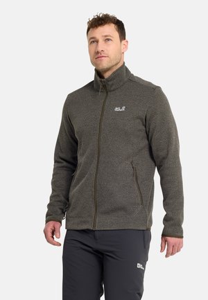 Mann trägt eine braune, strukturierte Jacke mit Reißverschluss und hohem Kragen sowie schwarze Hose, steht vor einem schlichten hellen Hintergrund.