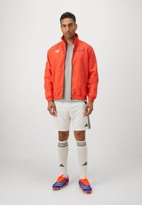 Röd vindjacka med hög krage, grå tröja, vita shorts och blå fotbollsskor med orange detaljer. Randiga vita strumpor.