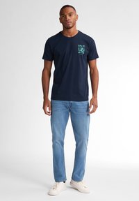 T-shirt en coton marine avec un graphique sur la poche avant, associée à un jean bleu clair et des baskets blanches, se tenant contre un fond clair.