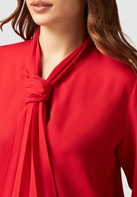 Blouse rouge à col noué, présentant un tissu drapé et une texture lisse. Le design comprend un nœud à l'avant et des bords plissés.