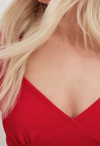 Rotes Stoffoberteil mit gerippter Textur, das über einen tiefen V-Ausschnitt und ein überkreuztes Design an der Brust verfügt. Akzente aus Stoff sammeln sich an der Taille.